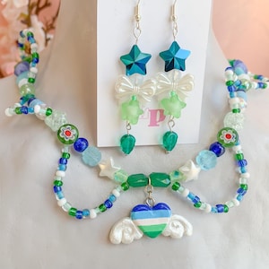 May include: A beaded necklace and matching earrings set. The necklace features a heart pendant with wings and a blue, green, and white color scheme. The earrings have star-shaped charms and bow accents. The jewelry set is in shades of blue, green, and white.