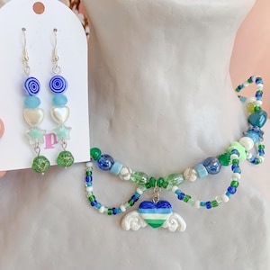 May include: A beaded necklace and matching earrings set. The necklace features blue, green, and white beads with a heart pendant and angel wings. The earrings have blue and green beads, hearts, and stars.