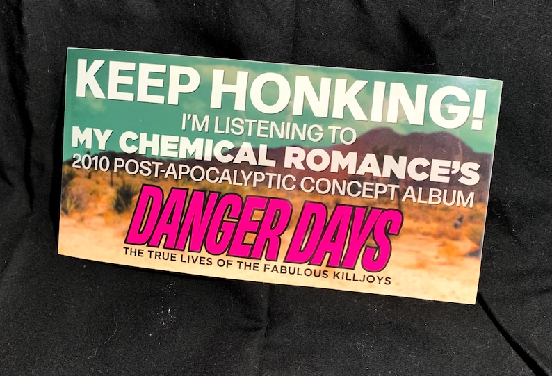 May include: Rectangular sticker with the text "KEEP HONKING! I'M LISTENING TO MY CHEMICAL ROMANCE'S 2010 POST-APOCALYPTIC CONCEPT ALBUM DANGER DAYS THE TRUE LIVES OF THE FABULOUS KILLJOYS" in white and pink lettering against a teal and desert landscape background.