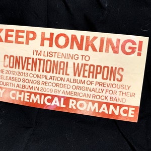 May include: A rectangular sticker with the text "KEEP HONKING! I'M LISTENING TO CONVENTIONAL WEAPONS" in red and black. The sticker also mentions "MY CHEMICAL ROMANCE" and details about an album.