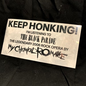 May include: A rectangular sticker with the text "KEEP HONKING! I'M LISTENING TO THE BLACK PARADE THE LEGENDARY 2006 ROCK OPERA BY MY CHEMICAL ROMANCE" in black font on a light gray background. The sticker is slightly angled.