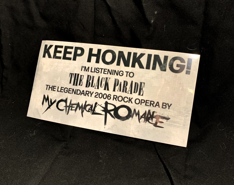 May include: A rectangular sticker with the text "KEEP HONKING! I'M LISTENING TO THE BLACK PARADE THE LEGENDARY 2006 ROCK OPERA BY MY CHEMICAL ROMANCE" in black font on a light gray background. The sticker is slightly angled.
