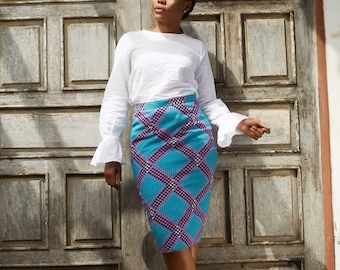 May include: A woman wearing a white long-sleeved blouse and a blue and pink patterned skirt. She is standing in front of a wooden door with a light brown background.