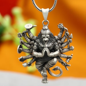 Op de afbeelding: Een zilveren ketting met een hanger met een gedetailleerde afbeelding van de hindoe-god Hanuman met meerdere armen die verschillende symbolen vasthouden. De hanger is bevestigd aan een zilveren ketting.