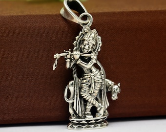 May include: Silver pendant featuring a detailed depiction of Krishna playing a flute, standing on a cow. The pendant has a loop at the top for attaching to a chain.