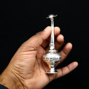 May include: A silver perfume bottle with intricate floral designs. The bottle has a long, slender neck and a small, round top.