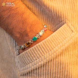 May include: A silver bracelet with seven oval-shaped gemstones. The gemstones are red, white, yellow, gray, green, and blue. The bracelet is on a person's wrist.