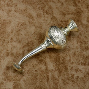 May include: A silver, ornate, decorative bottle with a long spout and a floral design. The bottle is lying on a brown, textured surface.