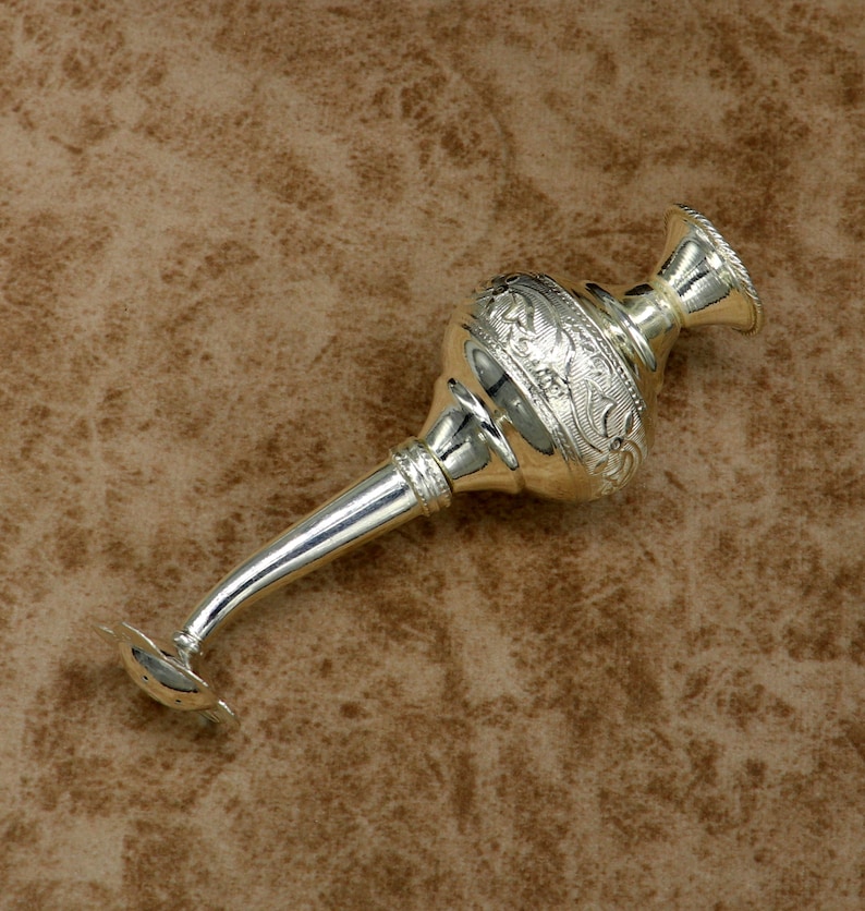 May include: A silver, ornate, decorative bottle with a long spout and a floral design. The bottle is lying on a brown, textured surface.