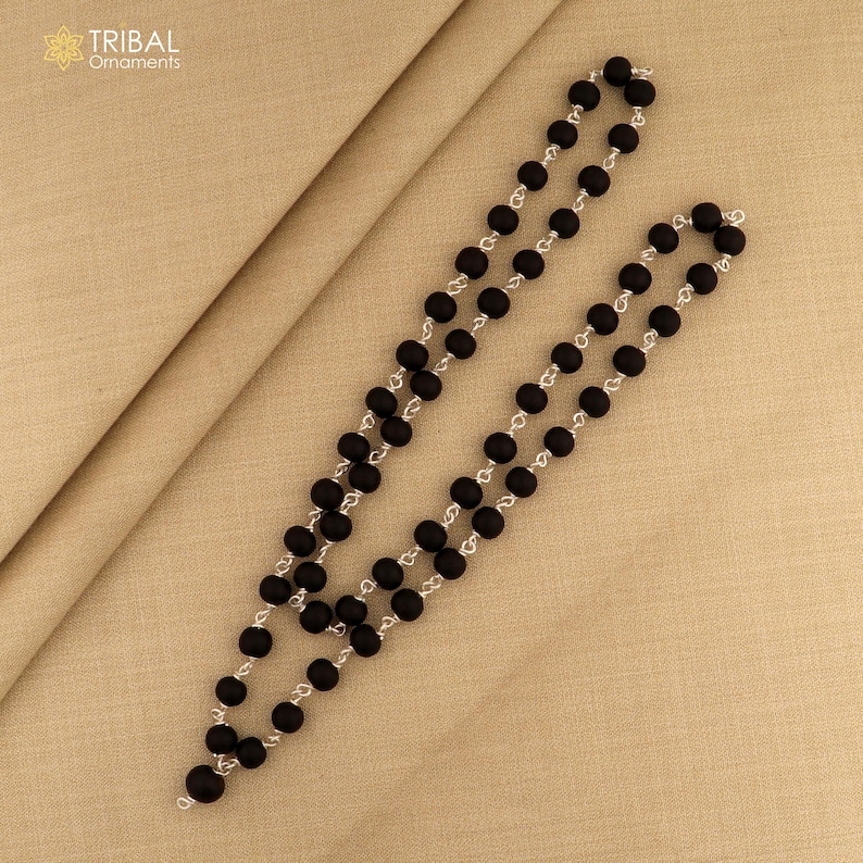 May include: Two strands of black beaded necklaces with silver-colored chain links, displayed on a beige fabric surface. The beads are round and evenly spaced. The necklaces are arranged diagonally across the frame.