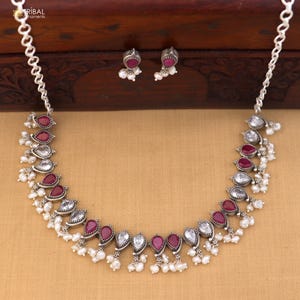 May include: A silver necklace and matching earrings set. The necklace features alternating teardrop-shaped red and clear stones, accented with small white pearls. The earrings mirror the design with red stones and pearls. The jewelry is displayed on a wooden surface.