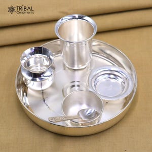 Könnte beinhalten: Ein silbernes Puja-Thali-Set mit mehreren Komponenten, darunter ein großer Teller, ein hoher Becher, eine kleine Schüssel, eine kleine Schale, ein kleiner Behälter und ein Löffel. Das Set besteht aus glänzendem Silbermetall und ist für religiöse Zeremonien bestimmt. Der Text "Tribal Ornaments" befindet sich oben links.