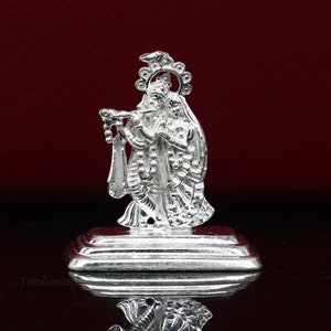 May include: A silver-colored statue of two figures, one playing a flute, on a rectangular base. The figures are adorned with detailed clothing and a halo. The statue is set against a dark red background.