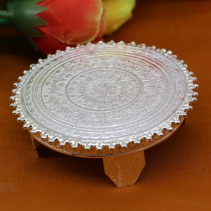 Puede incluir: Un soporte decorativo de plata con una parte superior redonda y tres patas. La parte superior presenta un intrincado diseño floral.