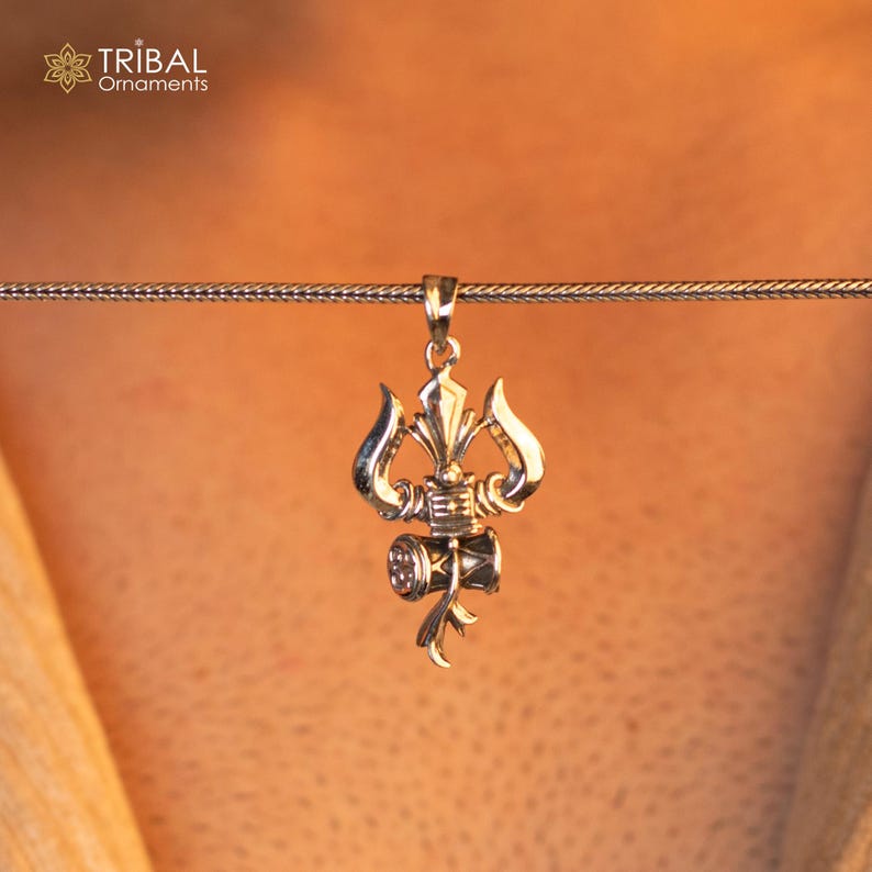 May include: Silver pendant necklace featuring a detailed design of a trident and a small drum, a symbol of the Hindu god Shiva.