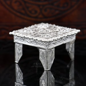 Puede incluir: Un soporte cuadrado decorativo de plata con intrincados diseños florales y bordes con cuentas. El soporte tiene cuatro patas con detalles ornamentados.