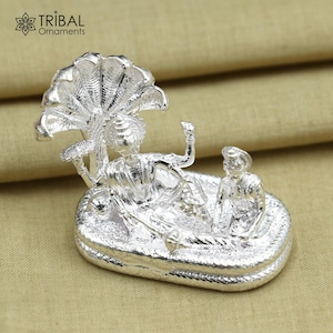 May include: A silver-colored religious figurine featuring two figures resting on a serpent-like base, with a multi-layered canopy overhead. The figurine has intricate details and is likely made of metal. The Tribal Ornaments logo is visible in the top left corner.