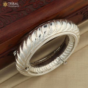 May include: A silver bangle bracelet with a decorative, textured design. The bracelet is open and has a hinged clasp.