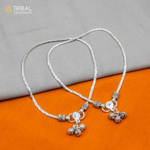 Könnte beinhalten: Zwei silberne Fußkettchen mit einem Kettenmuster und kleinen dekorativen Glöckchen an den Enden.