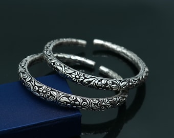 May include: Two silver-toned open-ended bangles featuring an intricate embossed floral and vine pattern throughout the surface. The metal bracelets have a darkened, oxidized finish in the recessed areas to highlight the detailed botanical engravings.