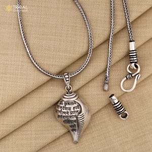 Op de afbeelding: Een zilveren ketting met een zilveren hanger in de vorm van een schelp. De hanger heeft Sanskriet-schriften erop. De ketting ligt op een bruine stoffen achtergrond.