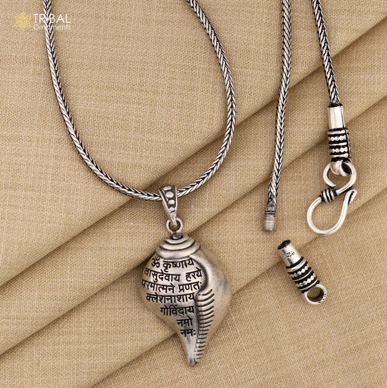 Op de afbeelding: Een zilveren ketting met een zilveren hanger in de vorm van een schelp. De hanger heeft Sanskriet-schriften erop. De ketting ligt op een bruine stoffen achtergrond.