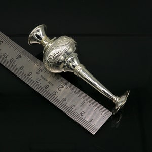 May include: A silver, ornate, decorative vase with a long, narrow spout. The vase is approximately 6 inches long and is resting on a ruler.