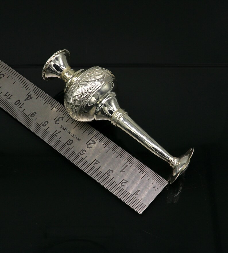 May include: A silver, ornate, decorative vase with a long, narrow spout. The vase is approximately 6 inches long and is resting on a ruler.
