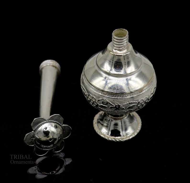 May include: Silver perfume bottle with a floral design and a separate nozzle. The bottle has a round body and a small, round base.