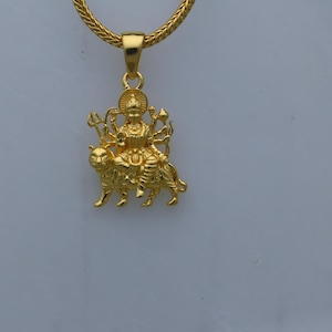 May include: A gold pendant necklace featuring a depiction of the Hindu goddess Durga riding a lion. The pendant is detailed with intricate carvings and a halo around the goddess's head.