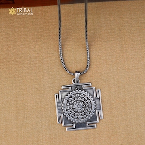 May include: A silver-colored pendant necklace featuring a square pendant with a detailed circular design at the center. The pendant is suspended from a matching silver chain. The brand name "Tribal Ornaments" is visible in the upper left corner.