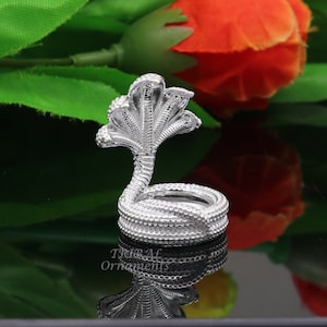 Peut inclure: Un porte-bague en forme de serpent argenté avec un design détaillé. Le serpent est enroulé, la tête levée et le capuchon déployé. Le porte-bague est posé sur une surface réfléchissante.