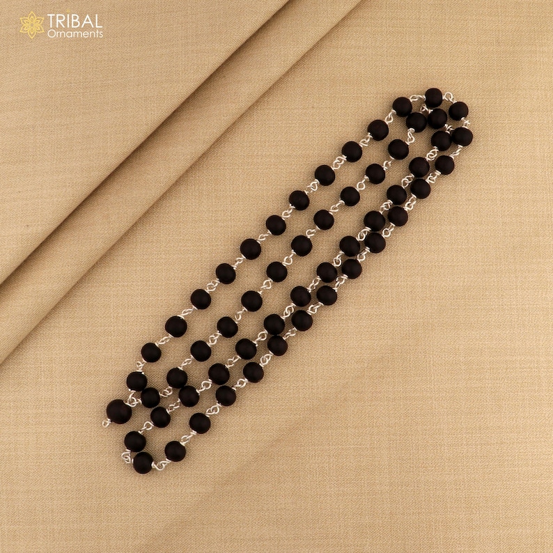 May include: Three strands of black beaded necklaces with silver-colored chain links, arranged diagonally on a beige fabric surface. The necklaces are composed of round, matte black beads. The upper left corner displays the text "TRIBAL Ornaments".