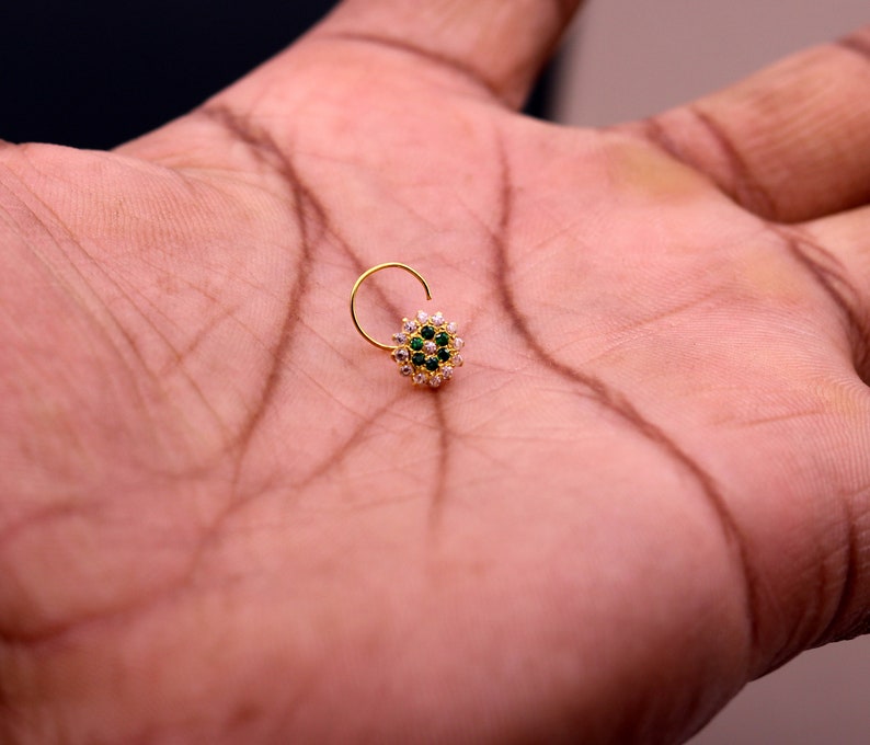May include: A gold nose ring with a circular design featuring white and green gemstones.