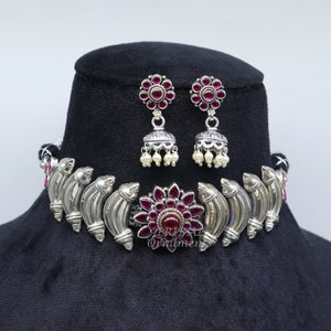 May include: Silver-toned choker necklace and matching earrings set. The necklace features a central floral design with a red gemstone and surrounding curved silver elements. The earrings have a floral design with red gemstones and dangling pearls.