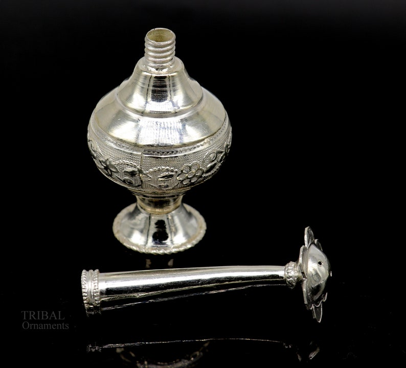May include: A silver, ornate, decorative bottle with a floral design and a separate nozzle. The bottle has a round body and a flared base. The nozzle is long and slender with a decorative top.