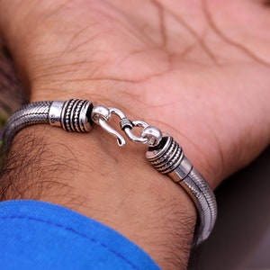 May include: A silver bracelet with a braided design and a hook clasp. The bracelet is on a person's wrist.