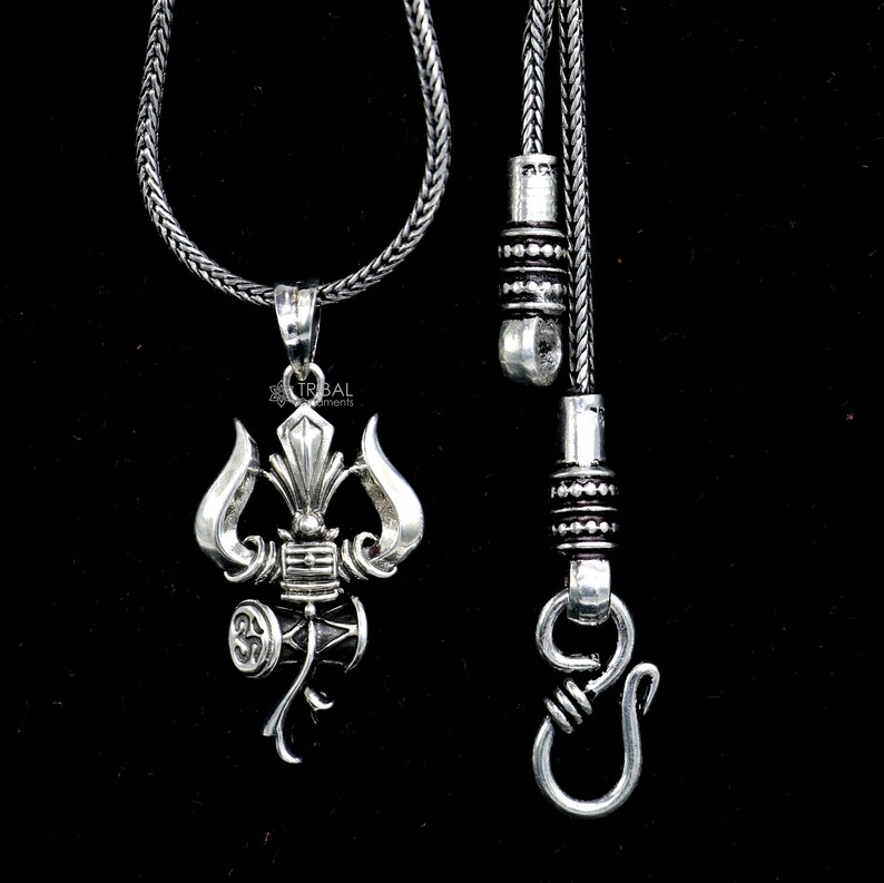 May include: A silver necklace with a pendant featuring a stylized depiction of a trident and a drum. The necklace has a silver chain and a silver clasp.