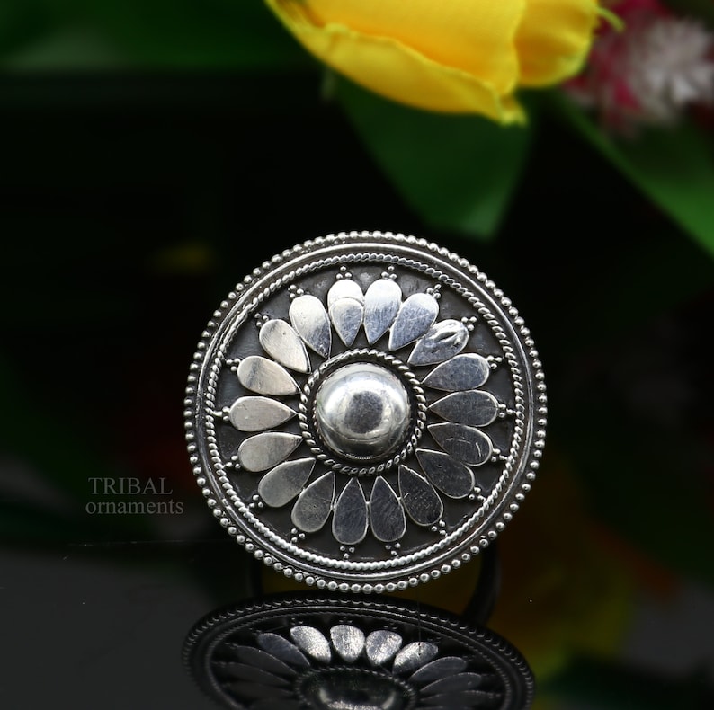 May include: A silver ring featuring a floral design. The ring is round and has a raised centre with a flower design. The flower has petals that are shaped like teardrops.