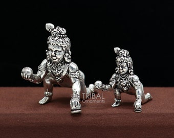 Könnte beinhalten: Zwei silberne Figuren von Baby Krishna, eine größer als die andere, beide krabbeln und halten eine Kugel. Die größere Figur ist im Vordergrund und die kleinere Figur im Hintergrund. Die Figuren sind detailliert und haben komplizierte Designs.