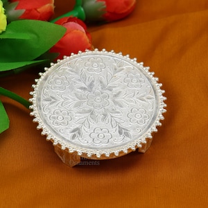 Puede incluir: Un soporte decorativo de plata con una parte superior redonda y tres patas. La parte superior presenta un intrincado diseño floral.