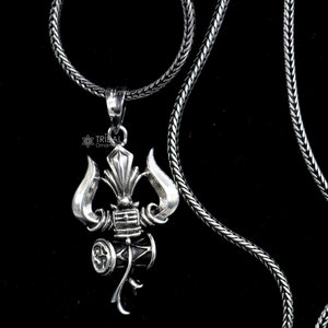 May include: A silver necklace with a detailed pendant featuring a trident and a drum. The pendant is a symbol of Shiva, a Hindu deity.