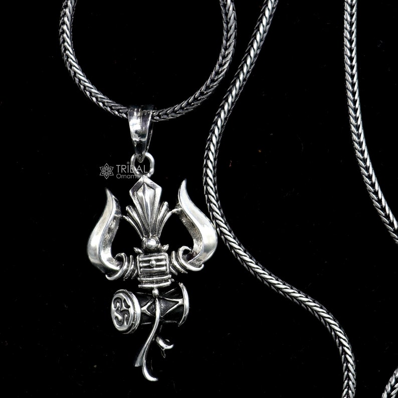 May include: A silver necklace with a detailed pendant featuring a trident and a drum. The pendant is a symbol of Shiva, a Hindu deity.