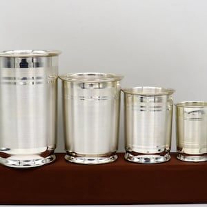 Op de afbeelding: Vier zilveren drinkglazen in verschillende maten, met een geborstelde afwerking en een decoratieve band rond het midden. De glazen zijn in een rij op een bruin oppervlak gerangschikt.
