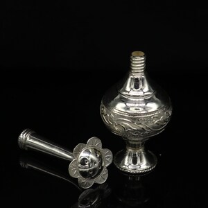 May include: A silver perfume bottle with an ornate design and a separate nozzle. The bottle has a round body and a short, wide neck. The nozzle is a long, thin tube with a flower-shaped end.