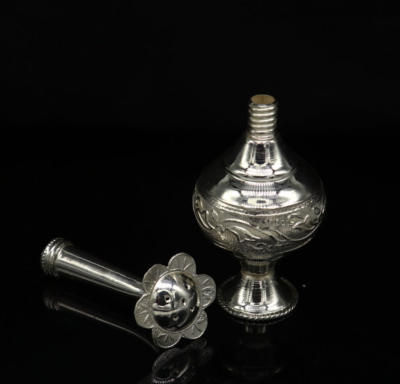 May include: A silver perfume bottle with an ornate design and a separate nozzle. The bottle has a round body and a short, wide neck. The nozzle is a long, thin tube with a flower-shaped end.