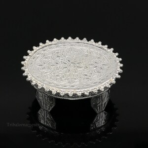 Puede incluir: Un soporte decorativo de plata con una parte superior redonda y tres patas. La parte superior tiene un diseño floral detallado.