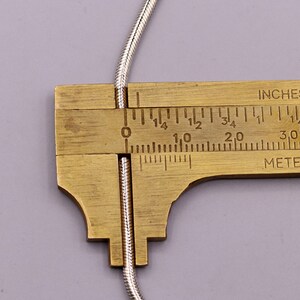 May include: A close-up of a silver snake chain being measured with a ruler. The ruler shows the chain is 2.5 cm long.