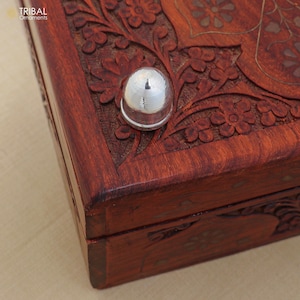 May include: Close-up of a handcrafted wooden box with intricate floral carvings. The box is stained a rich reddish-brown color and features a shiny silver metal knob. The text "TRIBAL Ornaments" is visible in the upper left corner.