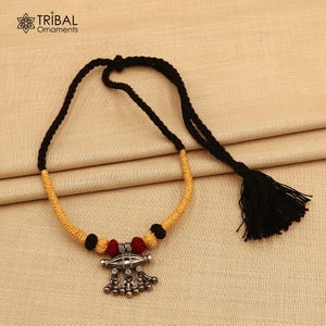 Puede incluir: Collar artesanal con un colgante plateado y cascabeles colgantes, cuentas rojas y negras, y un cordón amarillo y negro. El collar tiene una borla negra. La marca "Tribal Ornaments" está en la esquina superior izquierda.